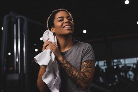 tattooed african american girl wiping sweat with towel and smiling n gymの写真素材
