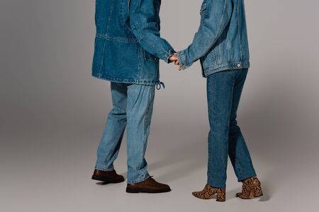 cropped view of stylish man and woman in denim jackets and jeans holding hands on grey background