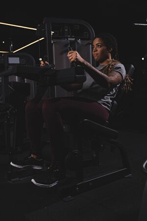 tattooed african american girl with dreadlocks working out on fitness machine