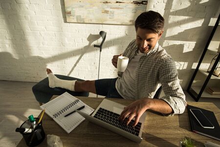 High angle view of smiling freelancer with broken leg holding cup and working at laptop at table