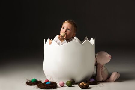 Child taking Easter egg to mouth by clenched hands inside eggshell on black background