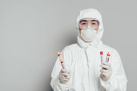 asian epidemiologist in hazmat suit and respirator mask holding test tubes with blood samples on grey backgroundの写真素材