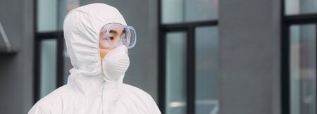 panoramic shot of asian epidemiologist in hazmat suit and respirator mask looking away while standing on street near buildingの写真素材