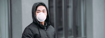 panoramic shot of young asian man in respirator mask looking away while standing on streetの写真素材