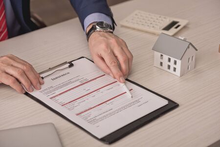cropped view of agent holding pen near clipboard with insurance lettering and carton house modelの写真素材
