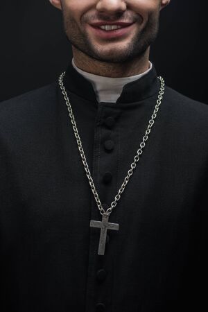cropped view of smiling catholic priest with silver cross on necklace isolated on blackの写真素材