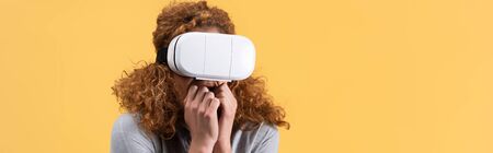 panoramic shot of scared redhead woman using vr headset, isolated on yellow