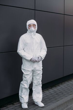asian epidemiologist in hazmat suit and respirator mask looking at camera while standing near wall