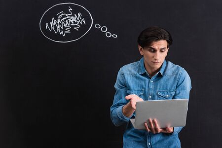 confused young man using laptop, with thought bubble drawing on blackboard