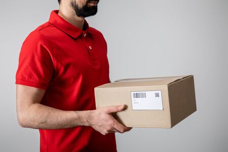 Cropped view of bearded courier in uniform carrying cardboard box isolated on greyの写真素材