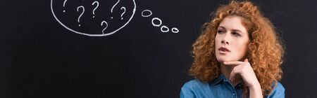 panoramic shot of beautiful thoughtful redhead girl with question marks in thought bubble on blackboardの写真素材