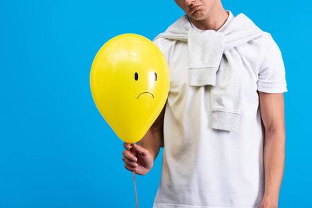 cropped view of sad man holding yellow upset balloon, isolated on blueの写真素材