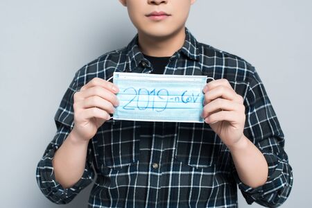 cropped view of young man holding respirator mask with 2019-ncov lettering on grey backgroundの写真素材