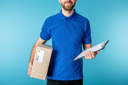 Cropped view of bearded courier holding clipboard and cardboard package isolated on blueの写真素材