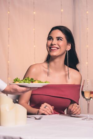 cropped view of waiter holding plate with fresh salad near cheerful, elegant girlの写真素材