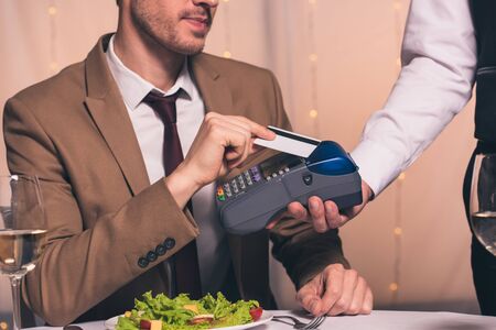 partial view of waiter with payment terminal near man holding credit card while sitting at served...
