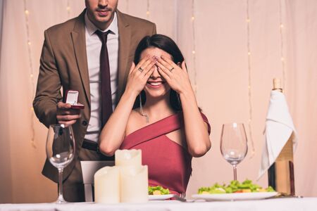 elegant man holding wedding ring near happy girlfriend sitting at served table and covering eyes with handsの写真素材