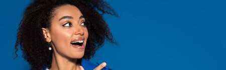 happy african american woman with open mouth pointing with finger aside isolated on blue, panoramic...