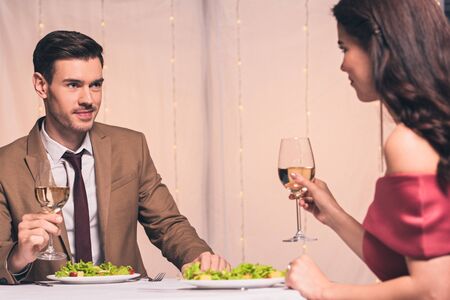 happy, elegant man and woman sitting at served table and holding glassed of white wineの写真素材