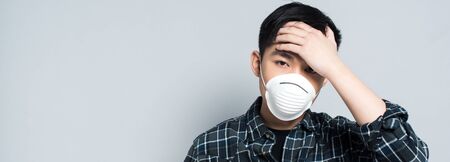 panoramic shot of young asian man in respirator mask touching forehead and looking at camera while...