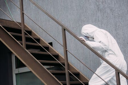 asian epidemiologist in hazmat suit and respirator mask walking upstairs outside building