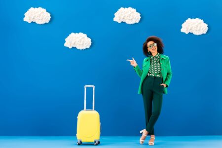 smiling african american woman pointing with finger and standing near travel bag on blue background with cloudsの写真素材