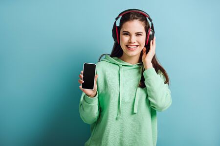 cheerful girl in wireless headphones looking at camera while showing smartphone with blank screen on...