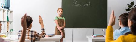 panoramic shot of schoolgirl standing near chalkboard with class work lettering and classmates with...