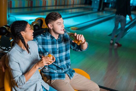 cheerful african american girl holding cocktail glass and talking to boyfriend drinking beer in bowling clubの写真素材