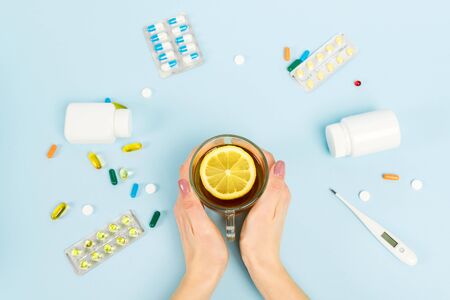 top view of woman holding cup of tea with sliced lemon near pills and digital thermometer on blueの写真素材