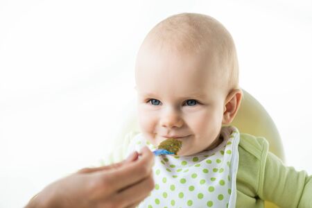 Selective focus of infant looking at mother with spoon while eating on feeding chair isolated on...