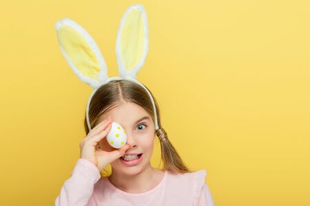emotional kid with bunny ears covering eye with dotted easter eggs isolated on yellow