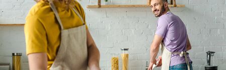 panoramic shot of happy bearded man looking at homosexual husband while washing plateの写真素材