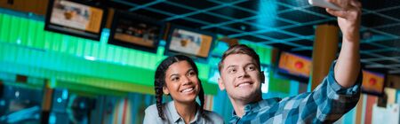 panoramic shot of happy interracial couple taking selfie on smartphone while sitting in bowling club