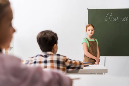 selective focus of upset schoolkgirl standing near chalkboard and classmatesの写真素材