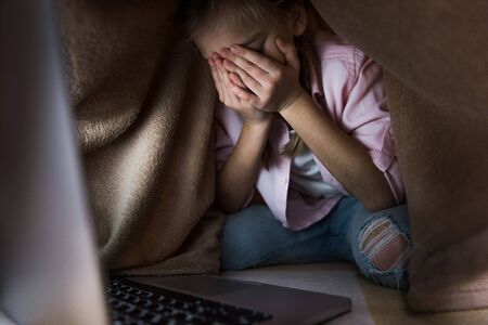 selective focus of frustrated kid covered in blanket covering face while crying near laptop,...