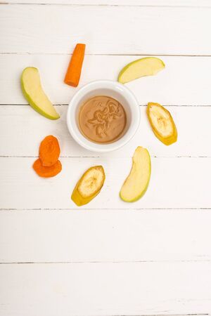 Top view of baby food in bowl with sliced fruits and carrot on white wooden surfaceの写真素材