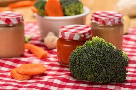 Close up view of fresh broccoli, carrot and cauliflower with jars of baby food on tablecloth on wooden surfaceの写真素材
