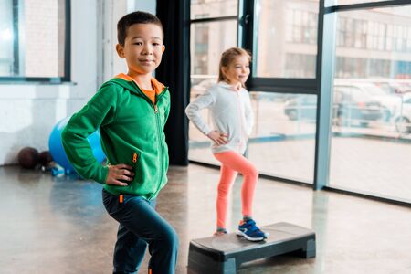 Selective focus of multicultural children with hands on hips doing exercise on step platforms in gymの写真素材