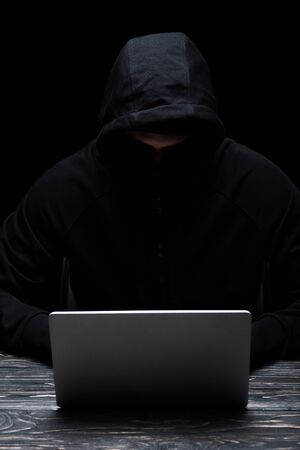 anonymous hacker in hood using laptop isolated on black