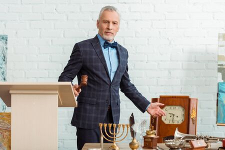 handsome auctioneer holding gavel and pointing with hand at antique objects during auction