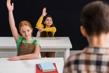 selective focus of schoolgirl with raised hands near classmate isolated on blackの写真素材
