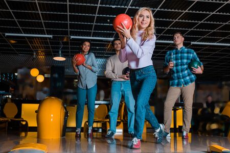 smiling blonde girl throwing bowling ball near multicultural friendsの写真素材