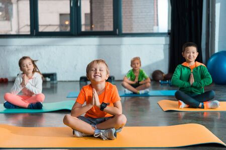 Selective focus of multiethnic children with clenched hands and crossed legs sitting on fitness matsの写真素材
