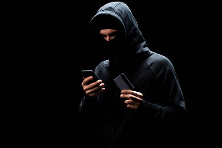 hacker in mask using smartphone and holding credit card isolated on black