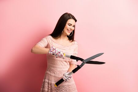 happy girl in working gloves holding gardening scissors on pink background