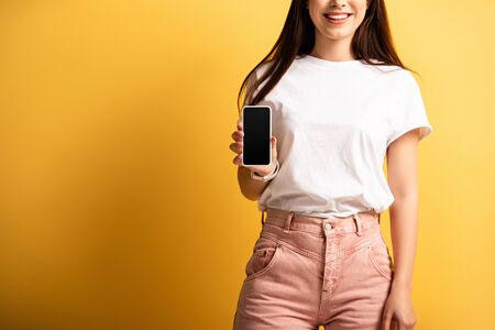 cropped view of smiling girl showing smartphone with blank screen on yellow background