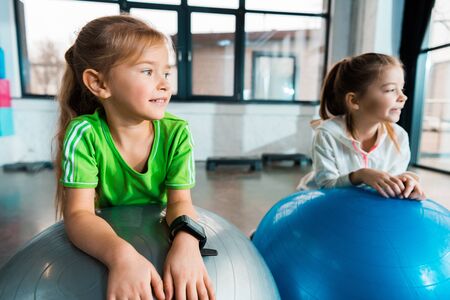 Selective focus of children looking away, smiling and leaning on fitness balls in gymの写真素材