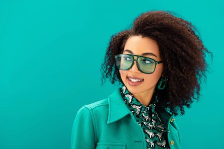 smiling african american woman in jacket looking away isolated on turquoiseの写真素材