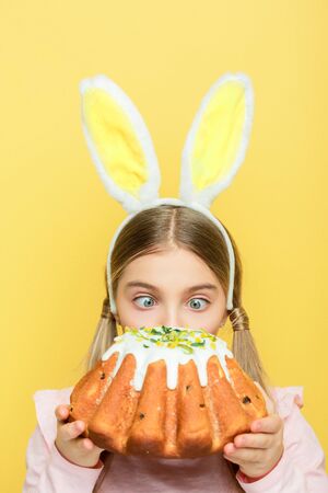 cute kid with bunny ears looking at easter cake isolated on yellow